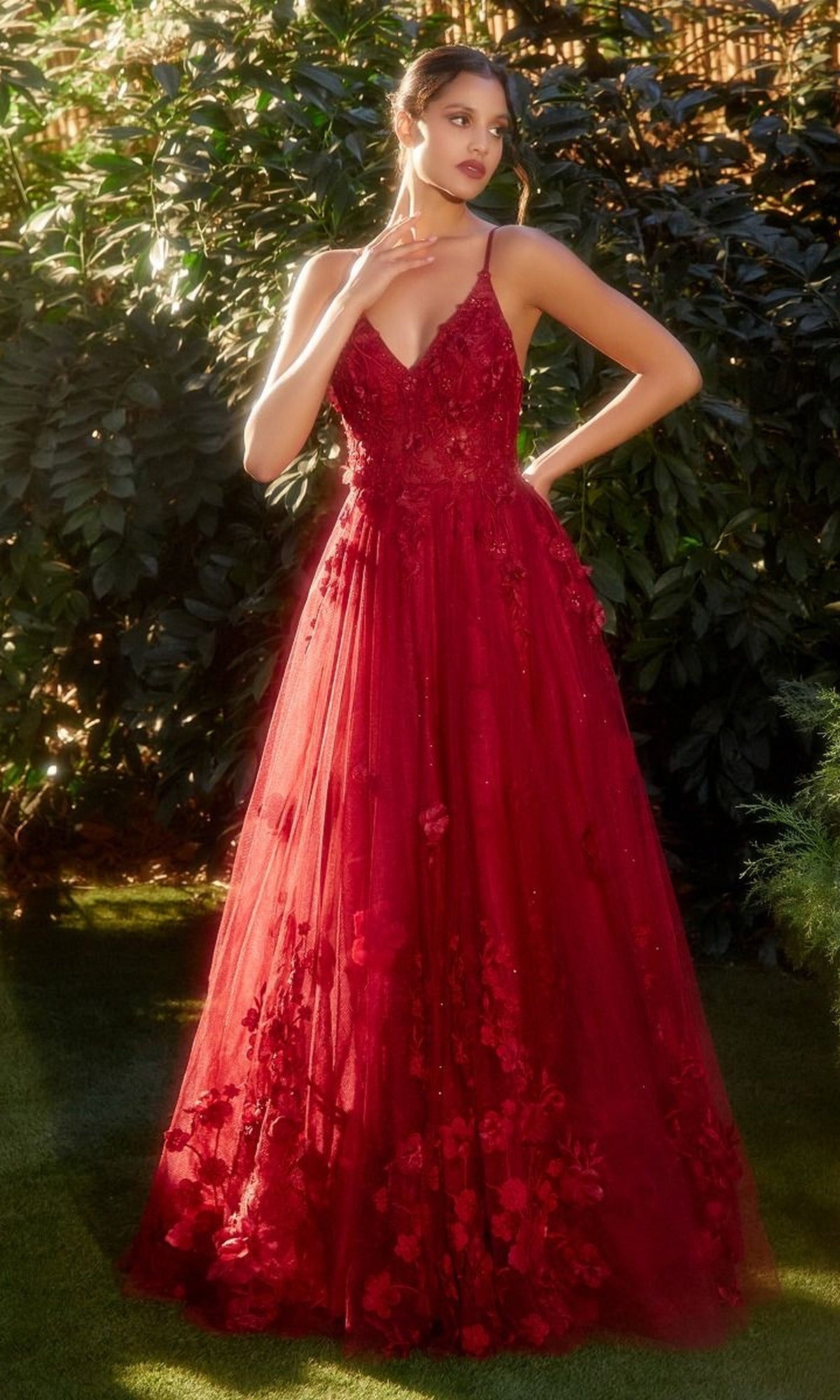 A woman stands outdoors in lush greenery wearing the Andrea and Leo A1326 Long Glitter Prom Ball Gown, a deep red, floor-length A-line dress with floral embellishments and thin straps.